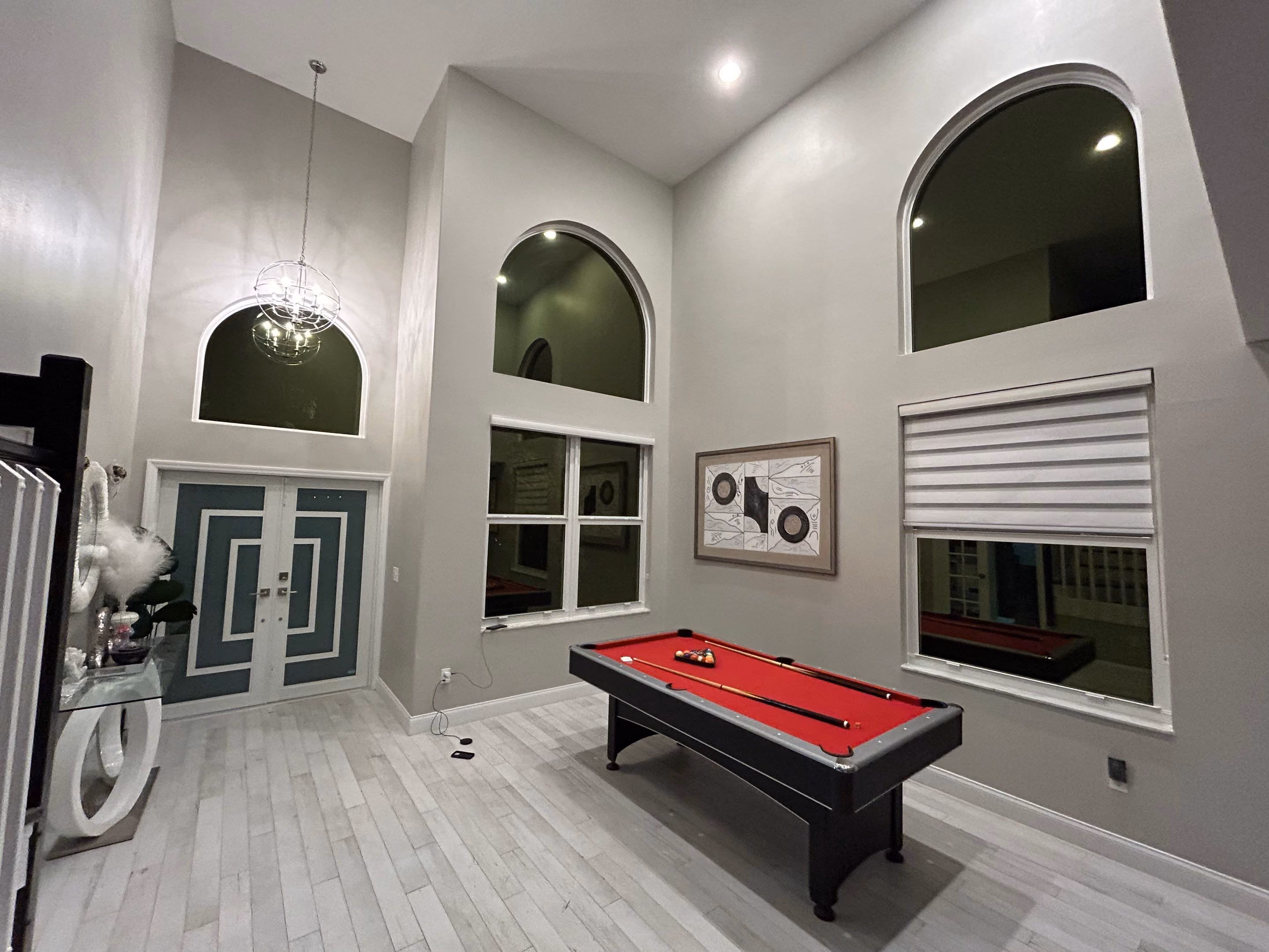 Modern game room with a red-felt pool table, geometric double doors, arched windows, and a framed abstract artwork on a light gray wall.