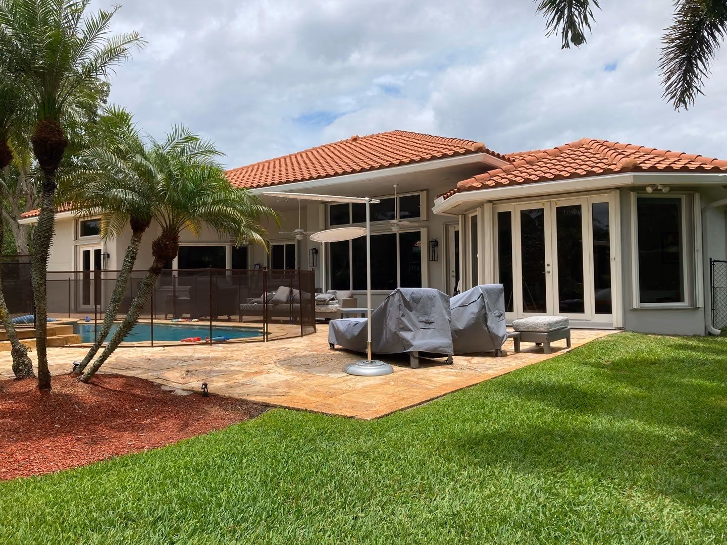 Backyard patio with covered outdoor chairs, green grass, palm trees, and a fenced swimming pool beside a house with a red tile roof.
