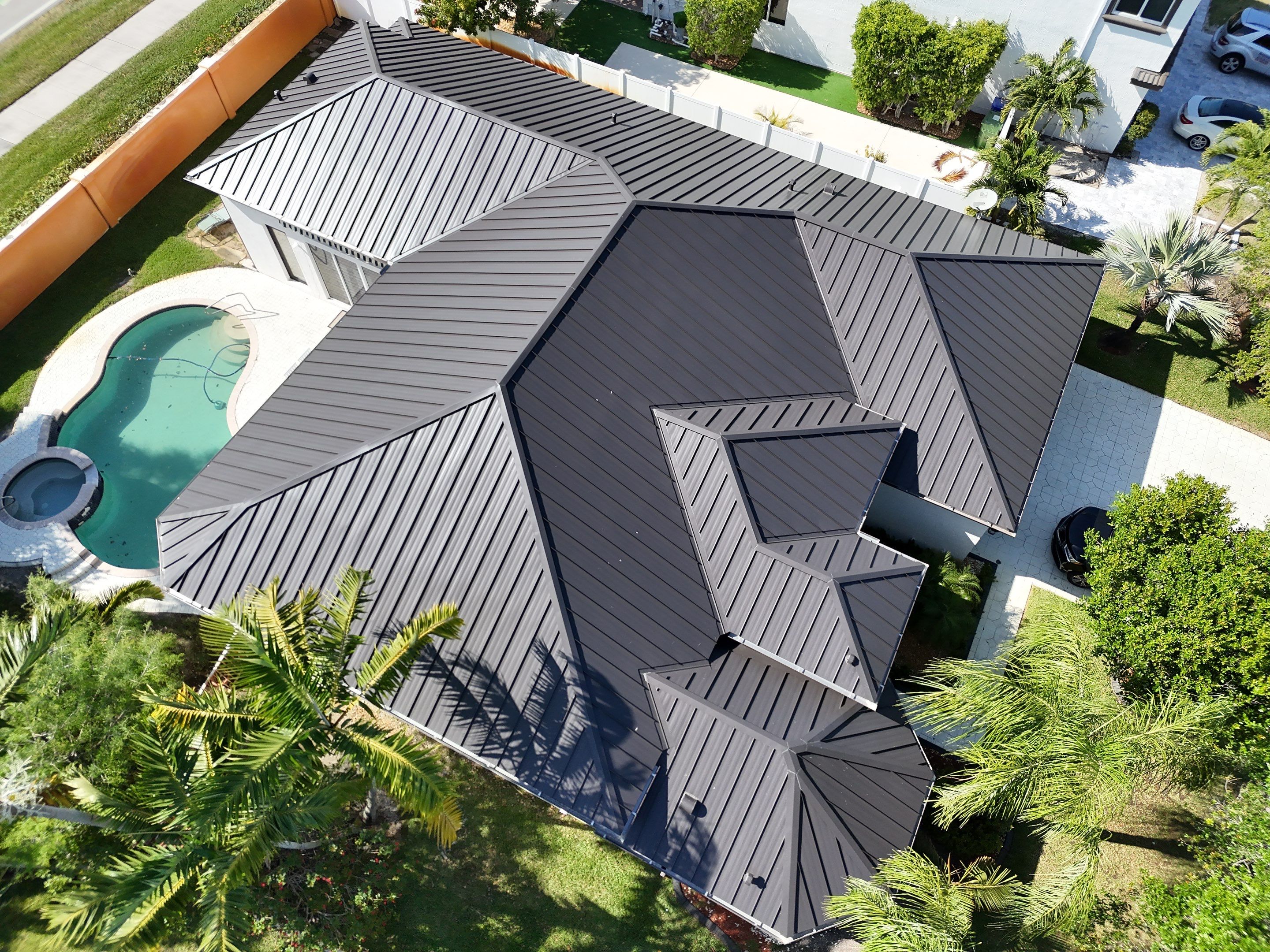 Aerial view of a modern house with a dark metal roof, a kidney-shaped swimming pool with an attached hot tub, surrounded by green palm trees and landscaped yard.