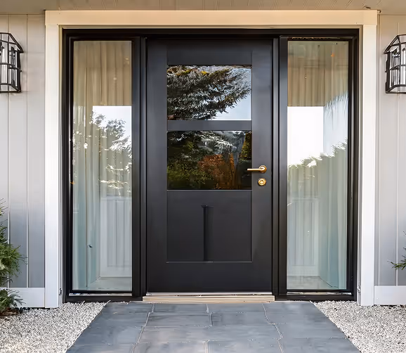 Modern black front door with glass panels on sides and brass handle, set in light-colored exterior wall with two wall lanterns.