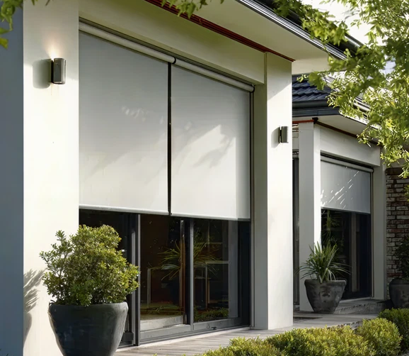Modern house exterior with large residential impact doors and white roller shades partially drawn, surrounded by potted plants and greenery.