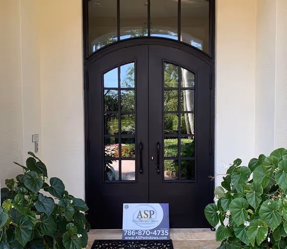 Black french front doors with glass panes reflecting greenery, flanked by two green leafy plants, and a sign for ASP Windows and Doors at the bottom.
