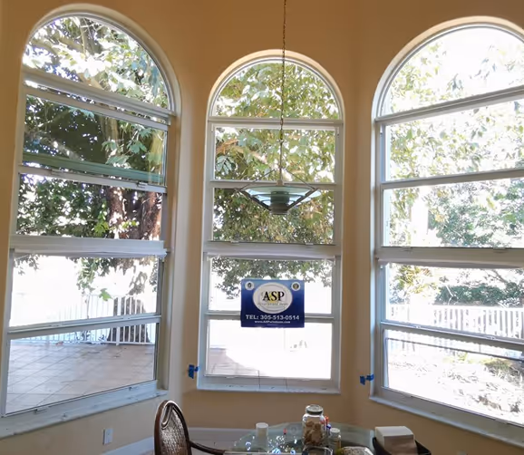 Three large residential impact windows in a room overlooking leafy trees outside during daytime.