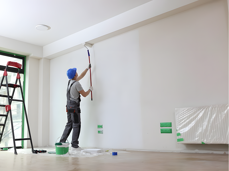 Worker wearing a blue helmet and gloves painting a white wall with a roller in a room under renovation.