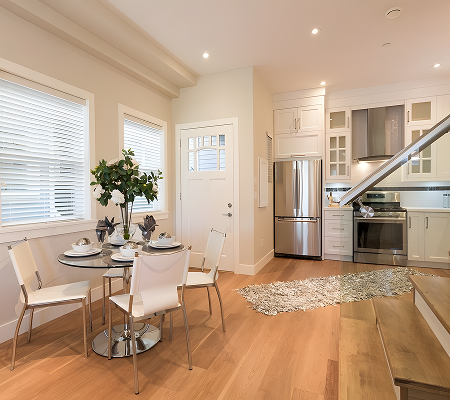 Interior decoration of modern kitchen and dining area with round glass table set for four, white chairs, stainless steel appliances, and wooden flooring.