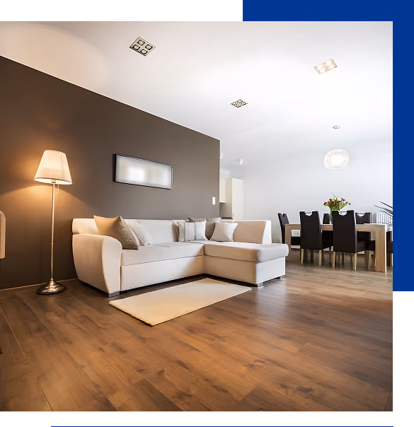 Modern living room with a beige sectional sofa, floor lamp, wall art, and wooden floor extending to a dining area with black chairs and a table with flowers.