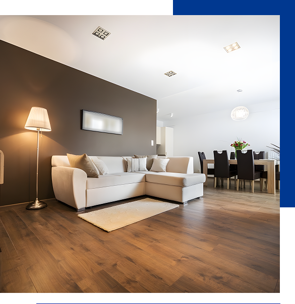 Modern living room with a beige sectional sofa, floor lamp, wall art, and wooden floor extending to a dining area with black chairs and a table with flowers.