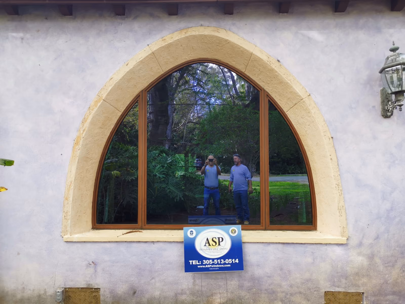 Arched window with wooden frames reflecting two men standing outside and greenery behind them, with an ASP Windows and Doors sign below the window.
