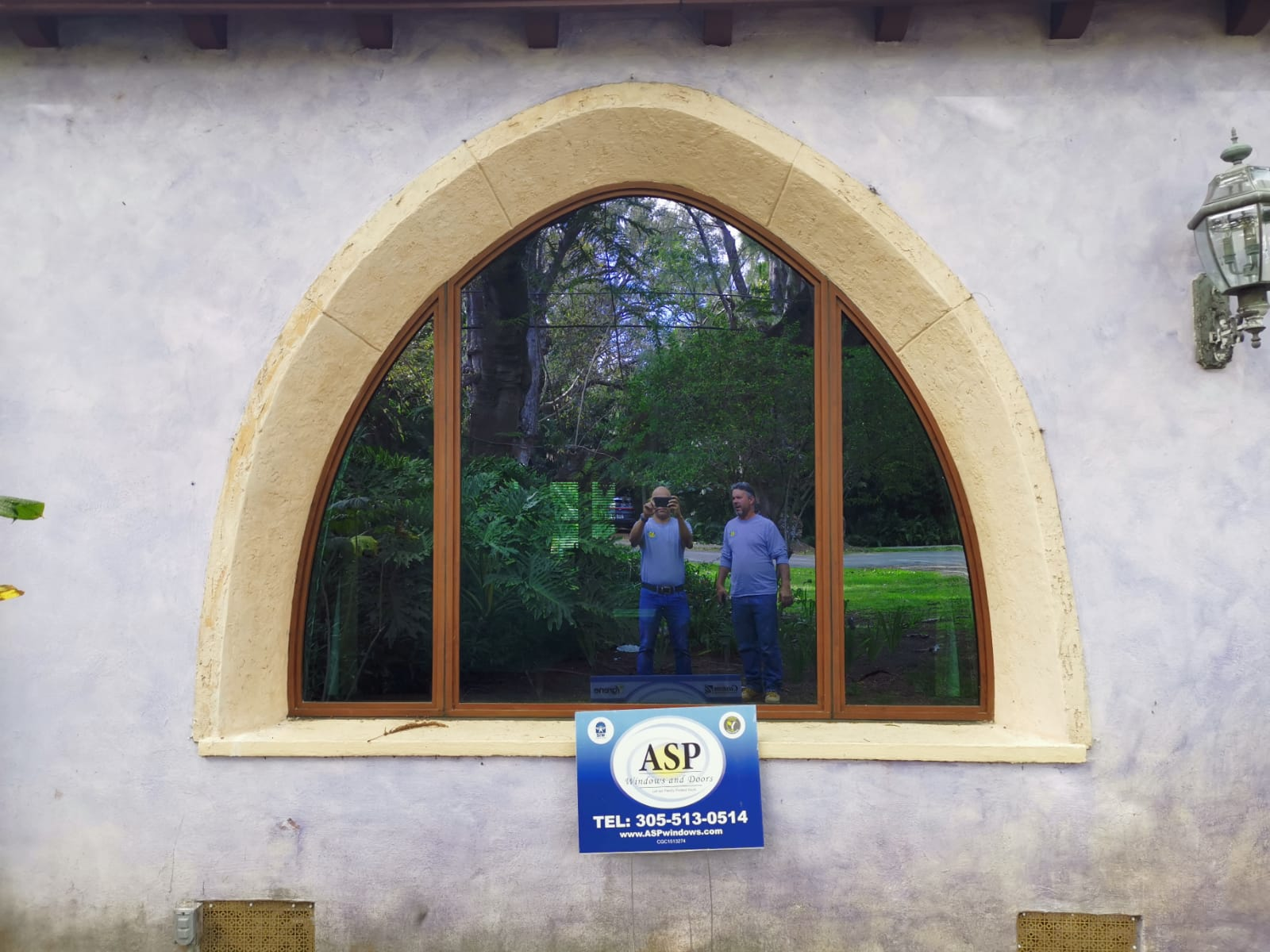 Arched window with wooden frames reflecting two men standing outside and greenery behind them, with an ASP Windows and Doors sign below the window.
