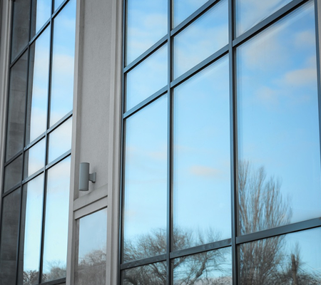 Reflection of a blue sky with clouds and tree branches on large glass windows of a building.