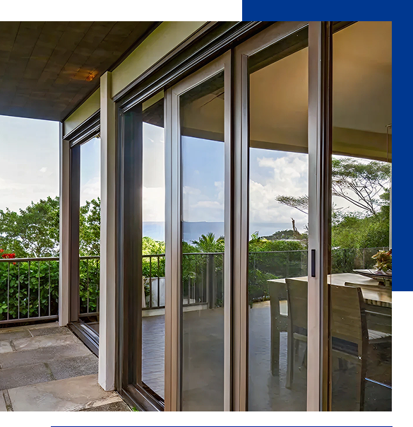 Sliding glass doors opening from a covered patio to a dining area with chairs, overlooking lush greenery and a distant ocean view.