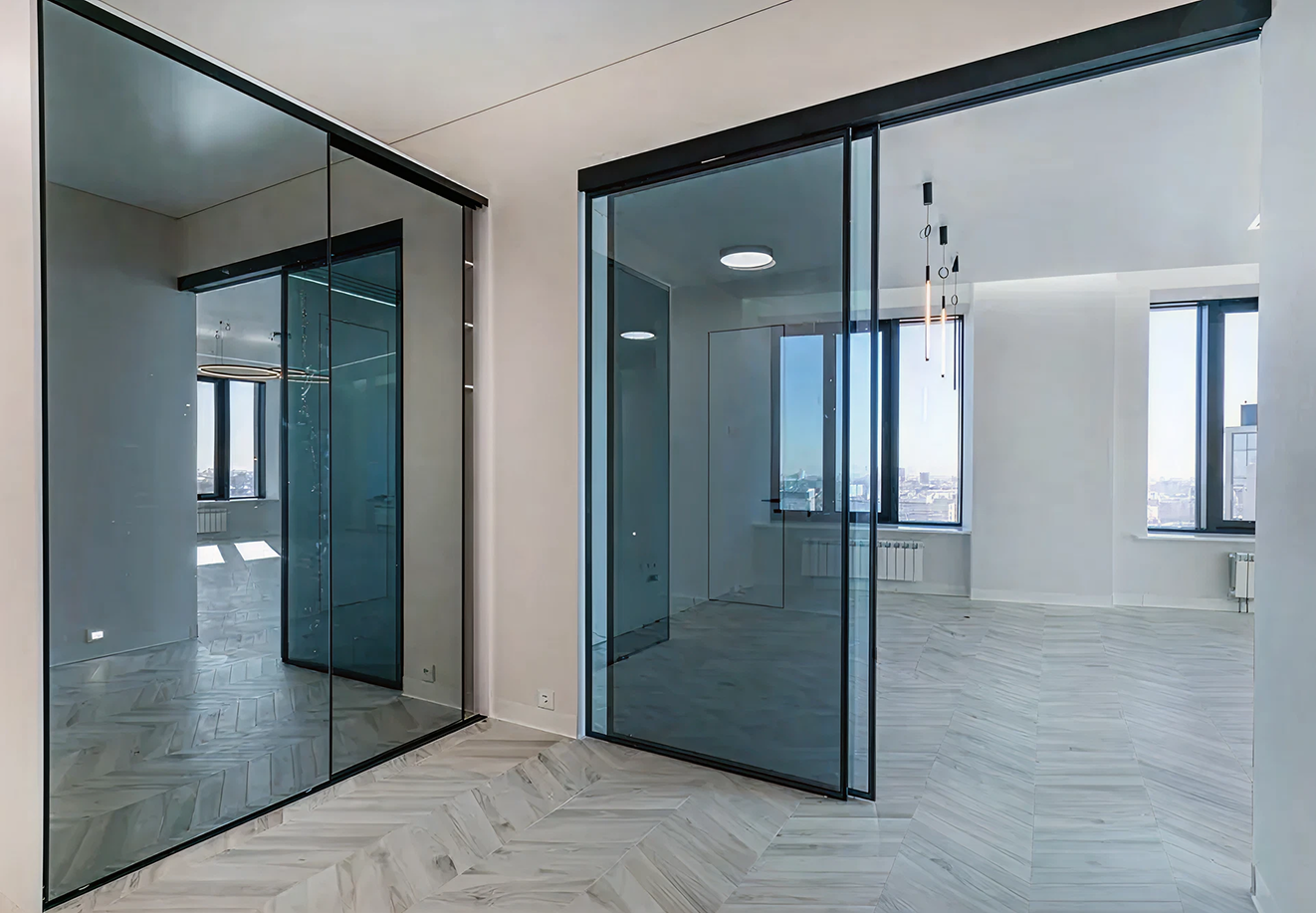 Modern empty room with large dark-framed glass sliding doors and light wood chevron flooring.