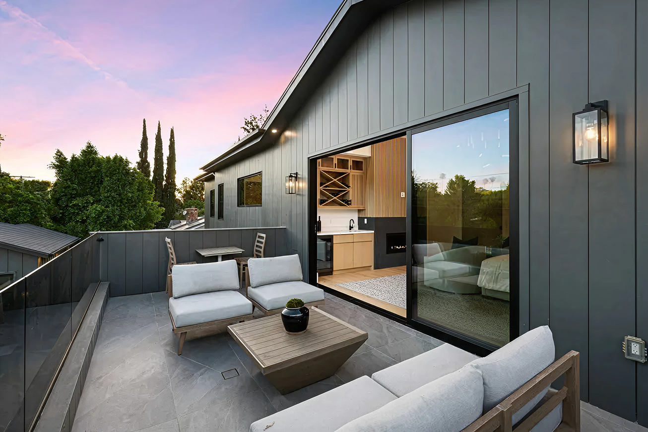 Modern outdoor patio with gray cushioned seating, wooden coffee table, and sliding glass door opening to a kitchen area at sunset.