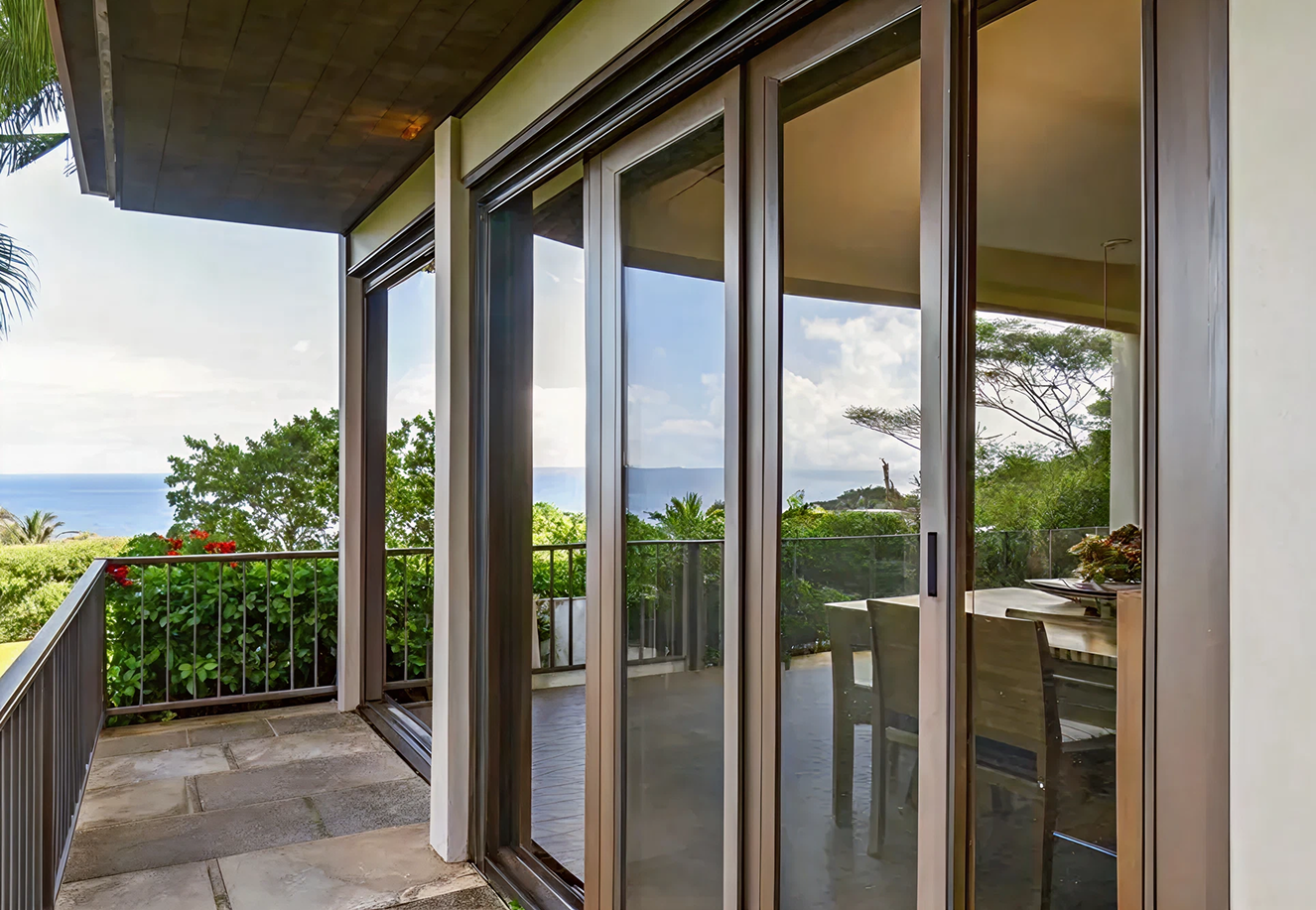 Balcony with stone floor and glass sliding doors reflecting a dining table and chairs, overlooking lush greenery and a distant ocean view.