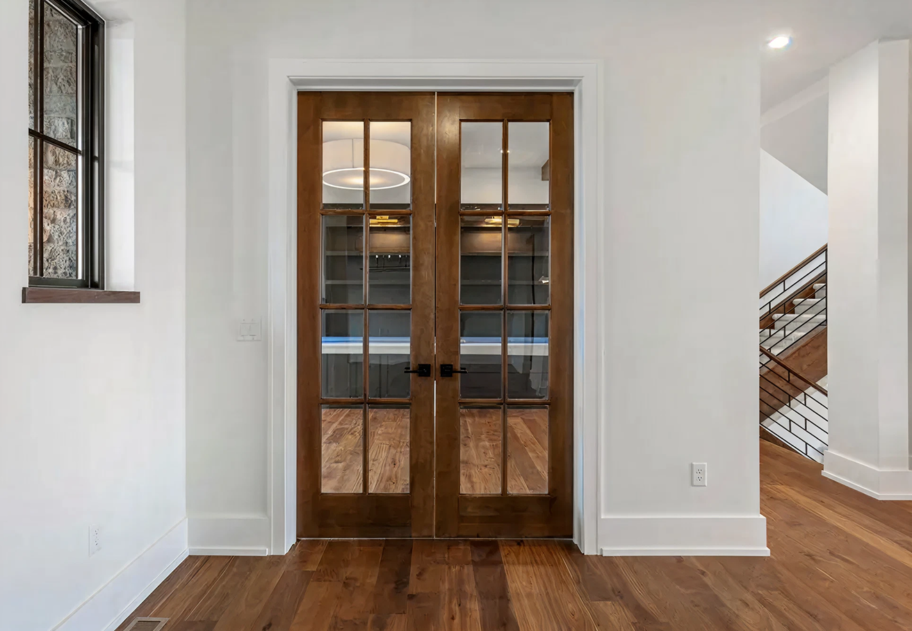 Wooden double french doors with glass panes leading to a room with hardwood floors and shelves.