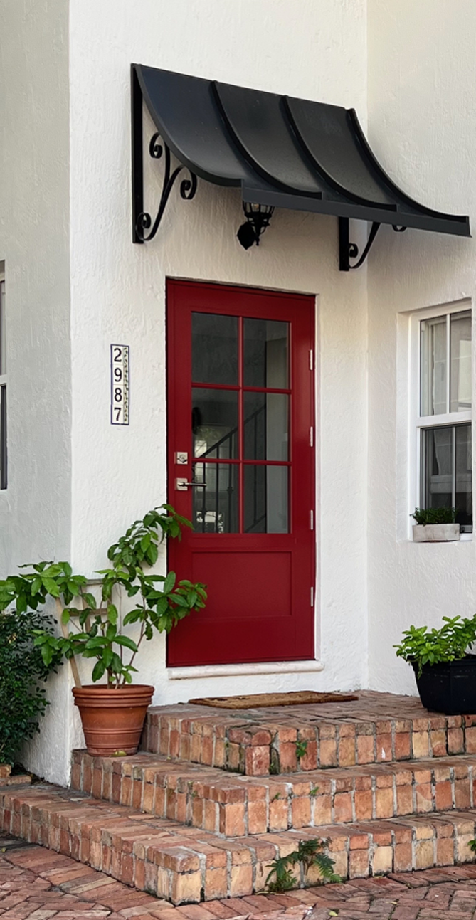 Red-framed entry door with a black curved metal awning above, set in a white stucco wall with brick steps and potted plants nearby.