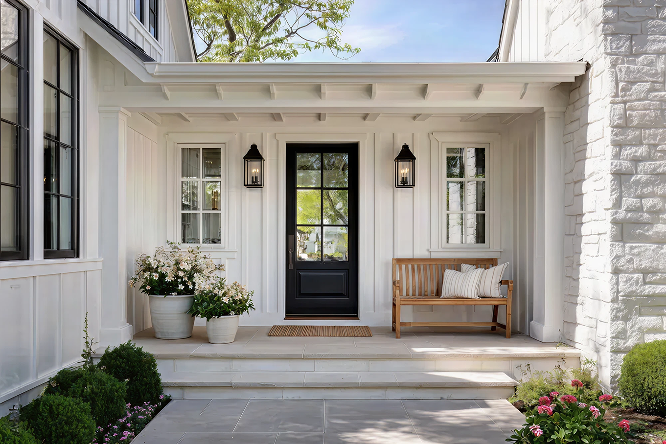 White front porch with entry door, two lantern-style wall lights, wooden bench with pillows, and potted plants.