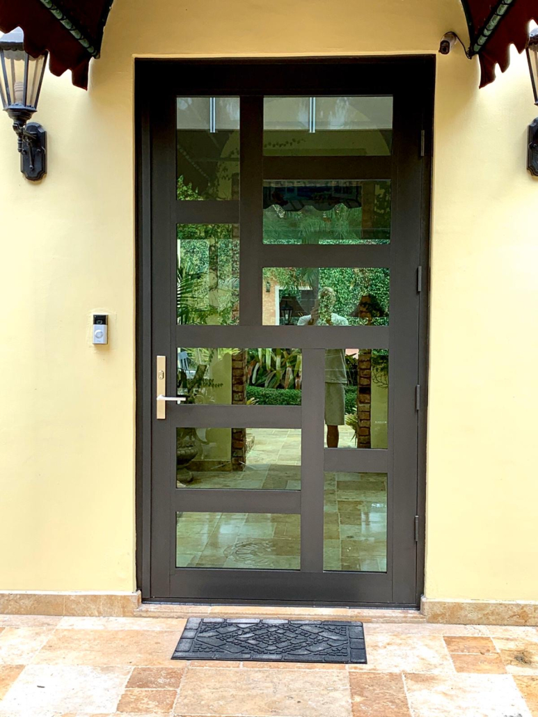 Modern dark glass entry door with geometric panels set in a yellow wall, flanked by two wall lamps and a doorbell, with a black decorative doormat on a tiled porch.