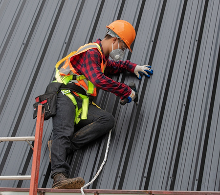 Construction worker wearing a safety helmet, mask, gloves, and harness, tightening screws on a metal roof