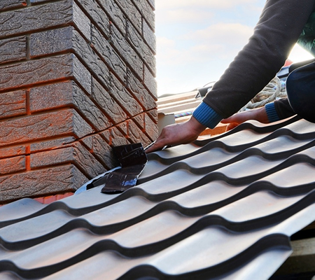 Person installing black metal roofing tiles near a brick chimney on a rooftop.