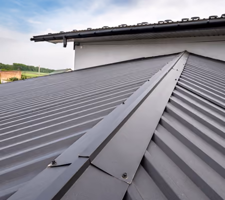 Close-up of a seam metal roof with overlapping panels and ridge cap under a partly cloudy sky.