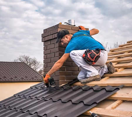 Worker in blue shirt and white pants installing black metal roofing panels near a brick chimney.
