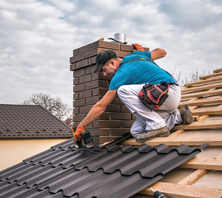 Worker in blue shirt and white pants installing black metal roofing panels near a brick chimney.