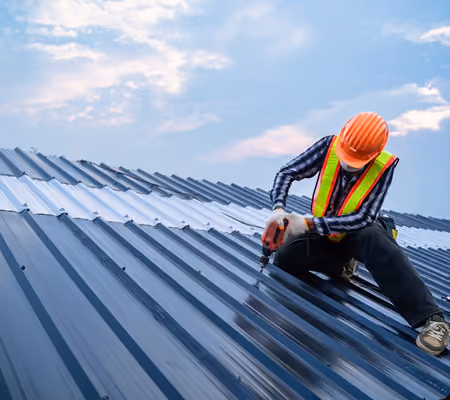 Construction worker in orange helmet and safety vest using power drill on metal roof against cloudy sky.