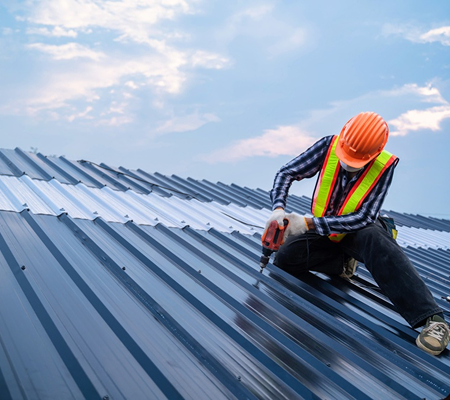 Construction worker in orange helmet and safety vest using power drill on metal roof against cloudy sky.