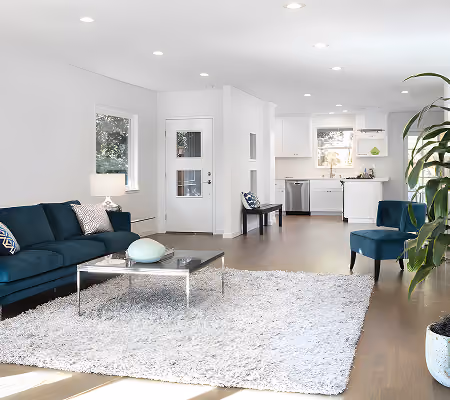 Bright living room with teal sofa and chair, white shag rug, glass coffee table, and view into a white kitchen.