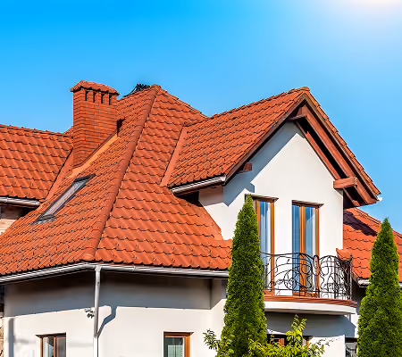 Modern house with red metal tiled roof, white walls, and two tall green shrubs in front under a clear blue sky.