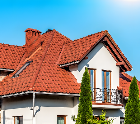 Modern house with red metal tiled roof, white walls, and two tall green shrubs in front under a clear blue sky.