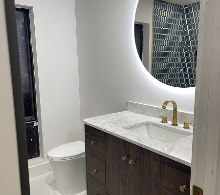 Modern bathroom with a white toilet, dark wood vanity with marble countertop, gold faucet, and illuminated round mirror reflecting a hexagonal tiled wall.