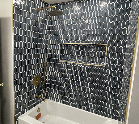 Bathtub with dark blue hexagonal tile walls and gold shower fixtures.