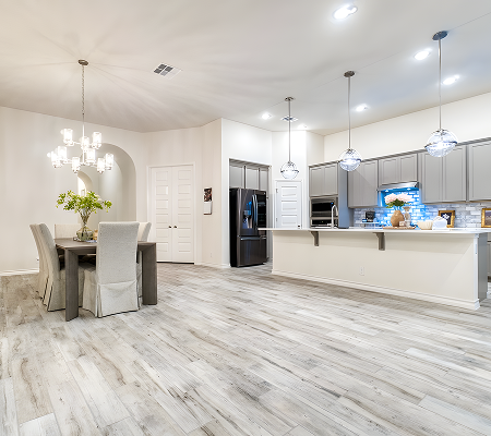 Bright, modern kitchen and dining area with light wood flooring, gray cabinets, black refrigerator, and a dining table with six upholstered chairs under a chandelier.