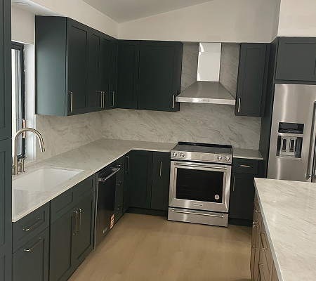 Modern kitchen with dark green cabinets, stainless steel oven, range hood, and refrigerator, white marble countertops, and a large window by the sink.