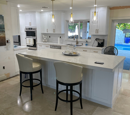 Bright modern kitchen with white cabinets, marble island, two beige bar stools, pendant lights, and a view of a backyard pool.