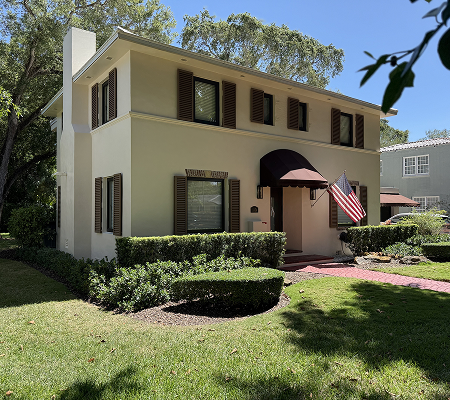 Two-story beige house with brown shutters, an American flag near the entrance, and a well-maintained front yard with green grass and bushes.