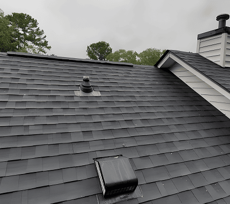 Dark gray asphalt metal tile roof with vents and chimney, trees visible in the background under a cloudy sky.