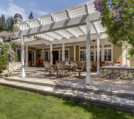 Backyard patio with a white pergola, outdoor dining table, chairs, and a garden area with stone borders and greenery.