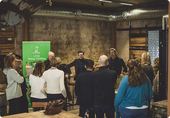 Group of people in casual attire standing and listening to two men speaking in an industrial-style room with exposed ducts and wooden walls.
