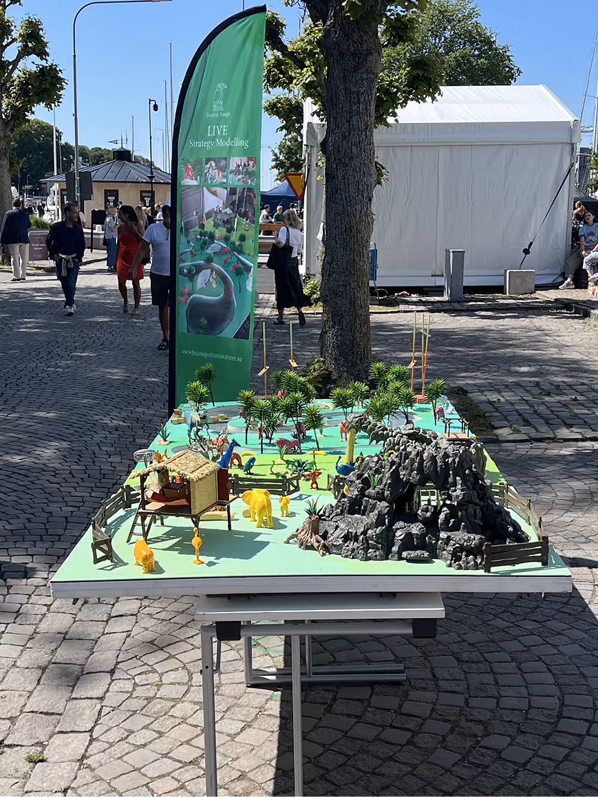 Outdoor display of a miniature wildlife model with trees, animals, and a rocky cave on a table, with a green banner behind it.