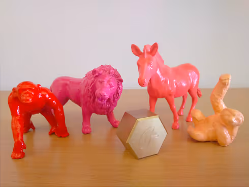 Brightly colored plastic figures of a red gorilla, pink lion, pink donkey, and an orange rabbit lying on its back on a wooden surface next to a metallic hexagonal object.
