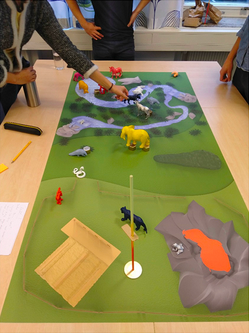 A tabletop diorama depicting a green landscape with toy animals including an elephant, cows, and birds, with several people interacting around it.