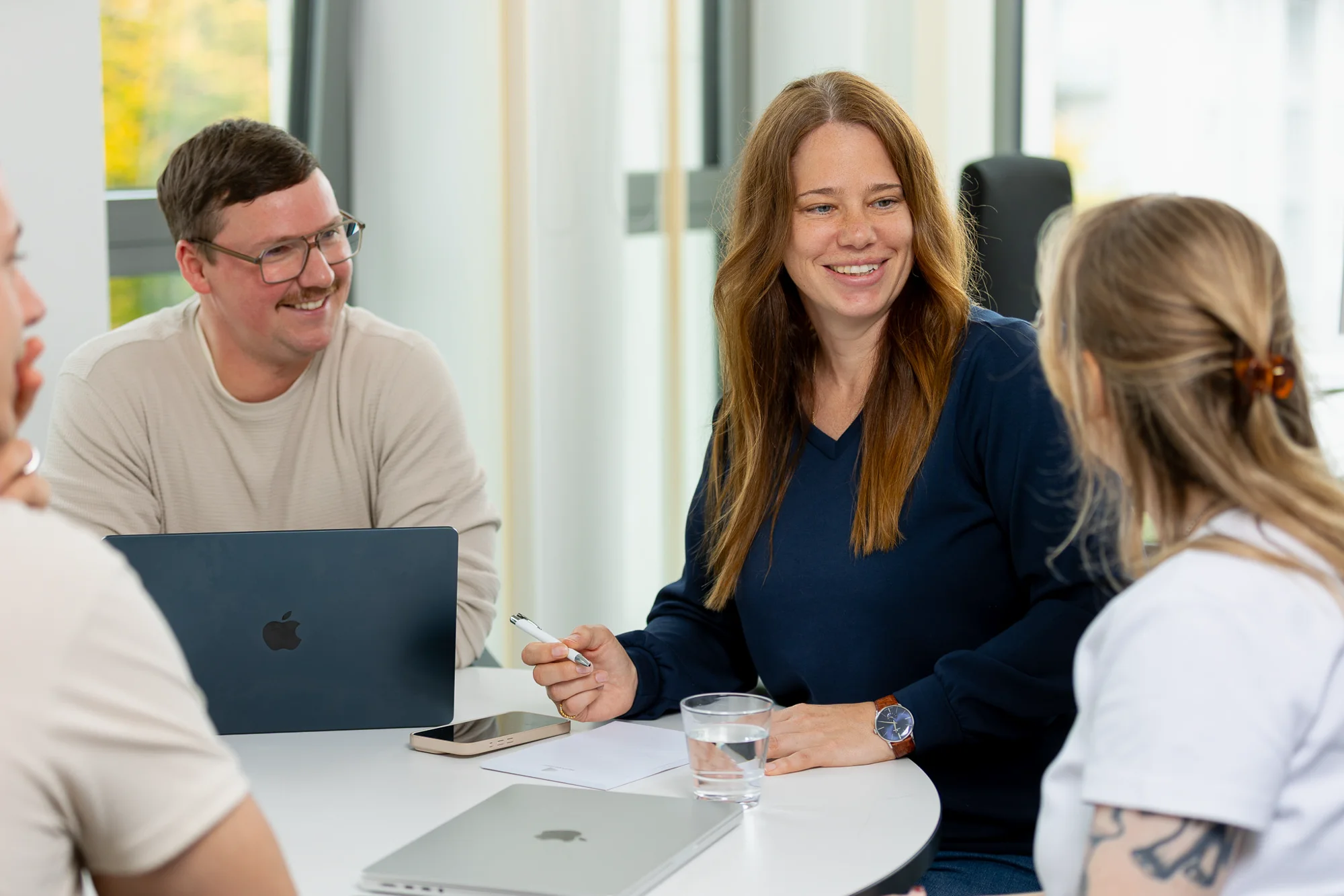 Vier Personen von 2BS sitzen an einem runden Tisch in einem hellen Büro und führen ein freundliches Gespräch. Auf dem Tisch befinden sich zwei Smartphone, ein Glas voll mit Wasser und zwei MacBooks, eines davon ist geöffnet und eines geschlossen.