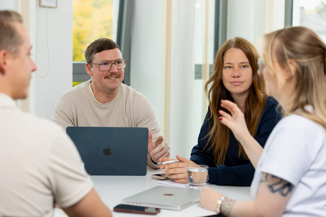Vier Personen von 2BS sitzen an einem Tisch und führen in einem hellen Büro eine Besprechung durch. Auf dem Tisch befinden sich zwei Smartphone, ein Glas voll mit Wasser und zwei MacBooks, eines davon ist geöffnet und eines geschlossen. Die Person ganz rechts im Bild gestikuliert mit ihrer rechten Hand.