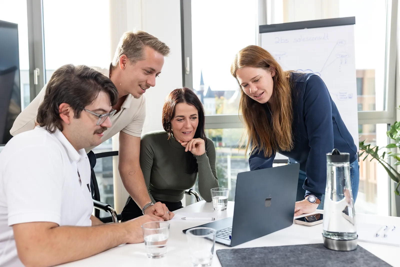 Vier Kolleginnen und Kollegen sitzen und stehen gemeinsam an einem Tisch und blicken auf einen Laptopbildschirm in einem hellen Büro.