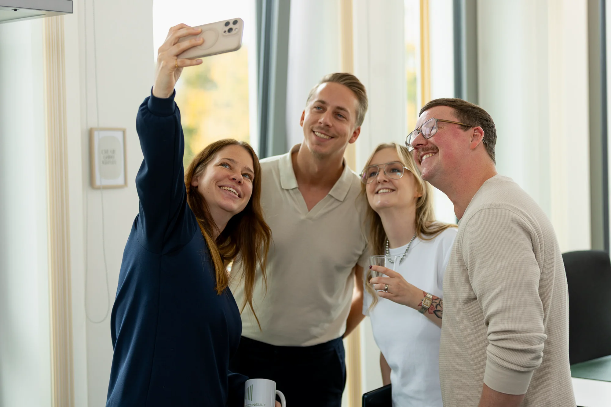 Vier Personen von 2BS machen zusammen ein Selfie in einem hellen Büro. Die Person ganz links macht das Selfie. Eine andere Person hat ein Glas in der Hand.