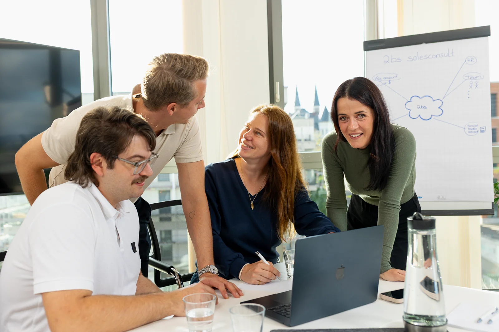 Vier Personen von 2BS arbeiten gemeinsam an einem MacBook in einem hellen Büro mit Flipchart und großen Bildschirm im Hintergrund.
