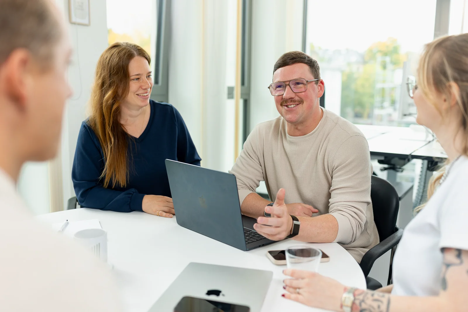 Vier Personen von 2BS sitzen an einem Tisch und führen in einem hellen Büro eine Besprechung durch. Auf dem Tisch befinden sich zwei Smartphone und zwei MacBooks, eines davon ist geöffnet und eines geschlossen. Die Person ganz rechts im Bild hat in  ihrer rechten Hand ein Glas.